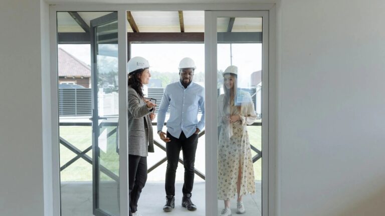 Three people wearing hard hats discussing construction plans near a sliding glass door.