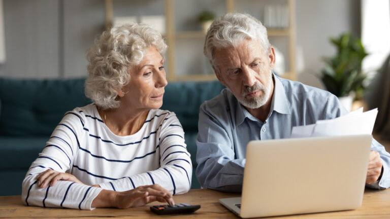 An older couple sitting at a table, engaged with a laptop.